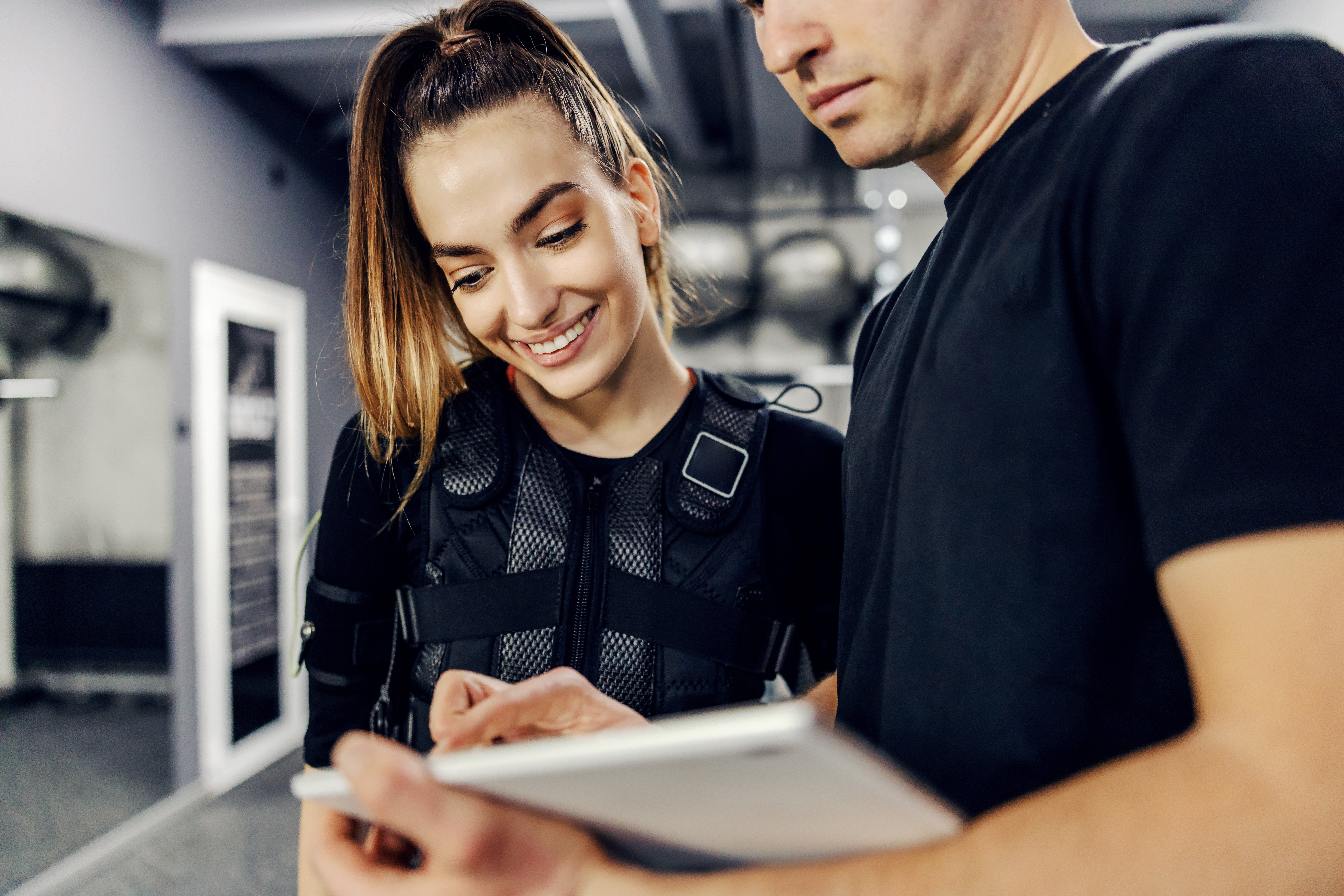 Satisfactory training results for electrical muscle stimulation training. Woman in a black EMS suit and a male trainer standing in the middle of the gym and observe the results on the tablet. Smile